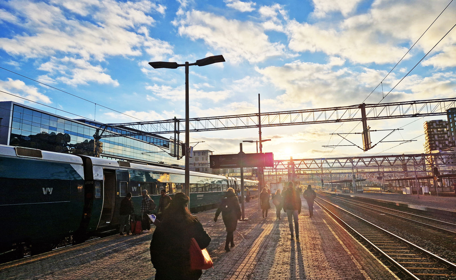 Lav sol over perrongene på Oslo S. Folk går på plattformen mot sola. Et tog står på ett av sporene.