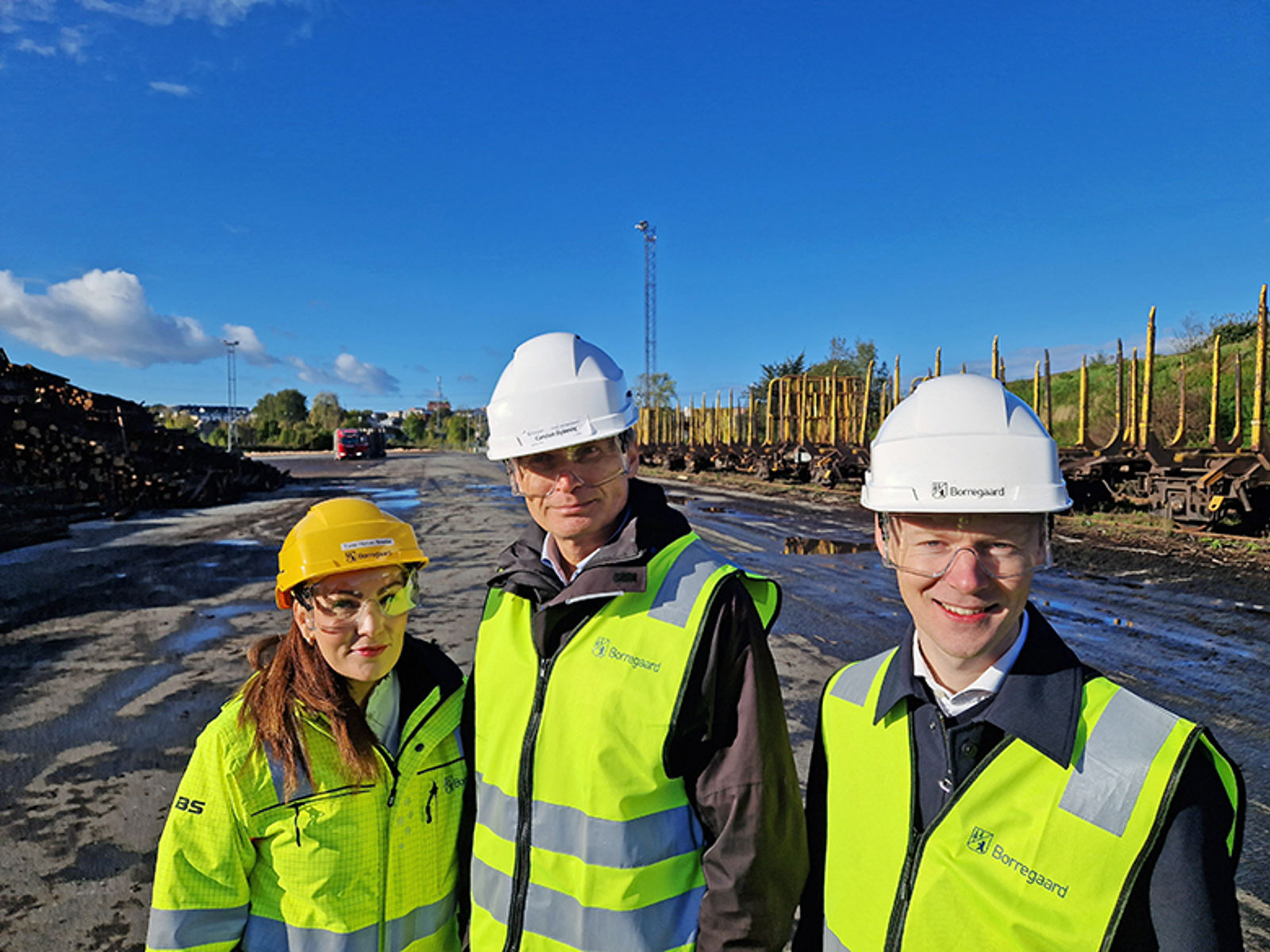 En kvinne og to menn i gule refleksvester og hjelmer, i et område med tømmerlagring og transport.