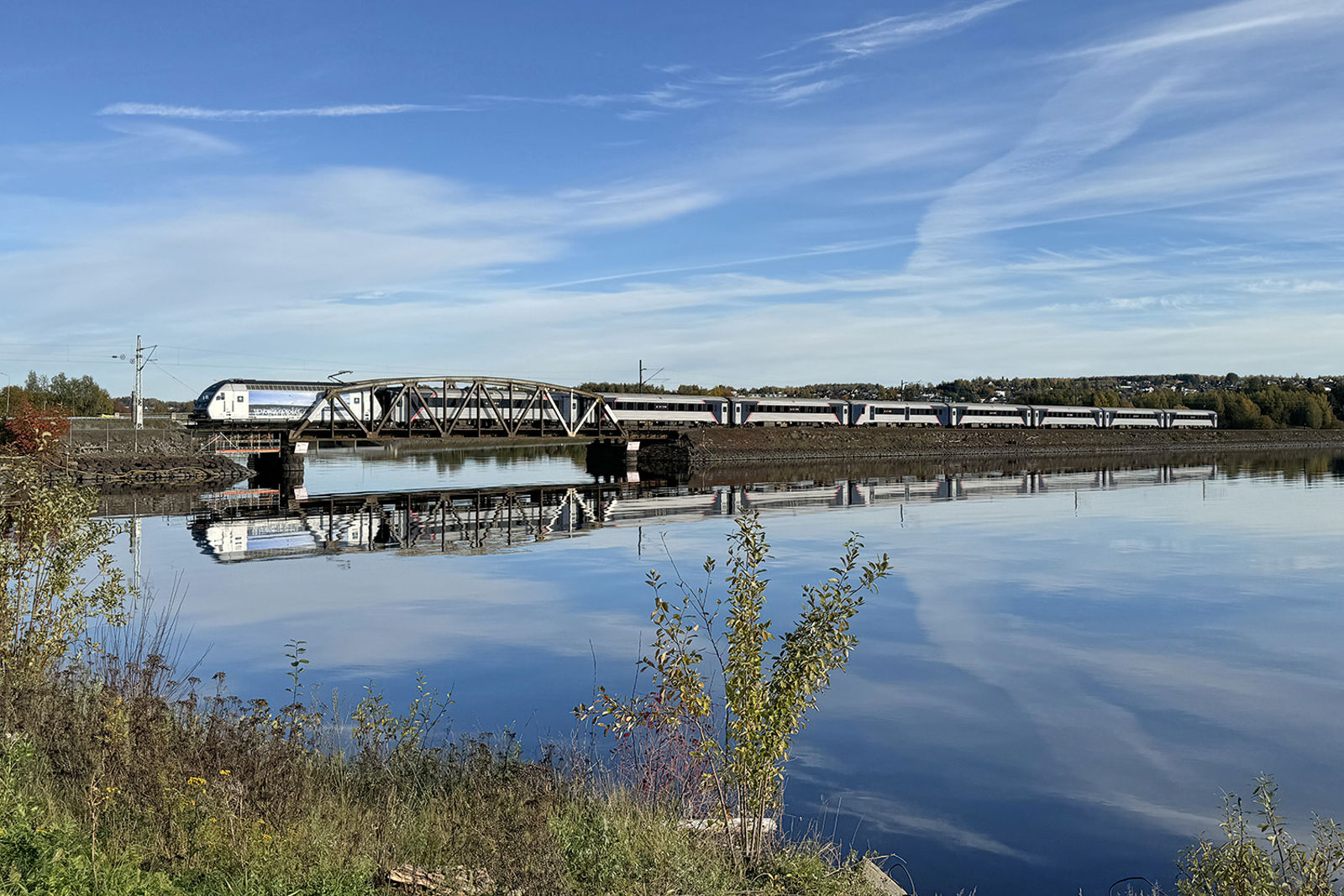 Høstbilde av tog på dagens enkeltspor med bru og fylling, toget speiler seg i vannet.