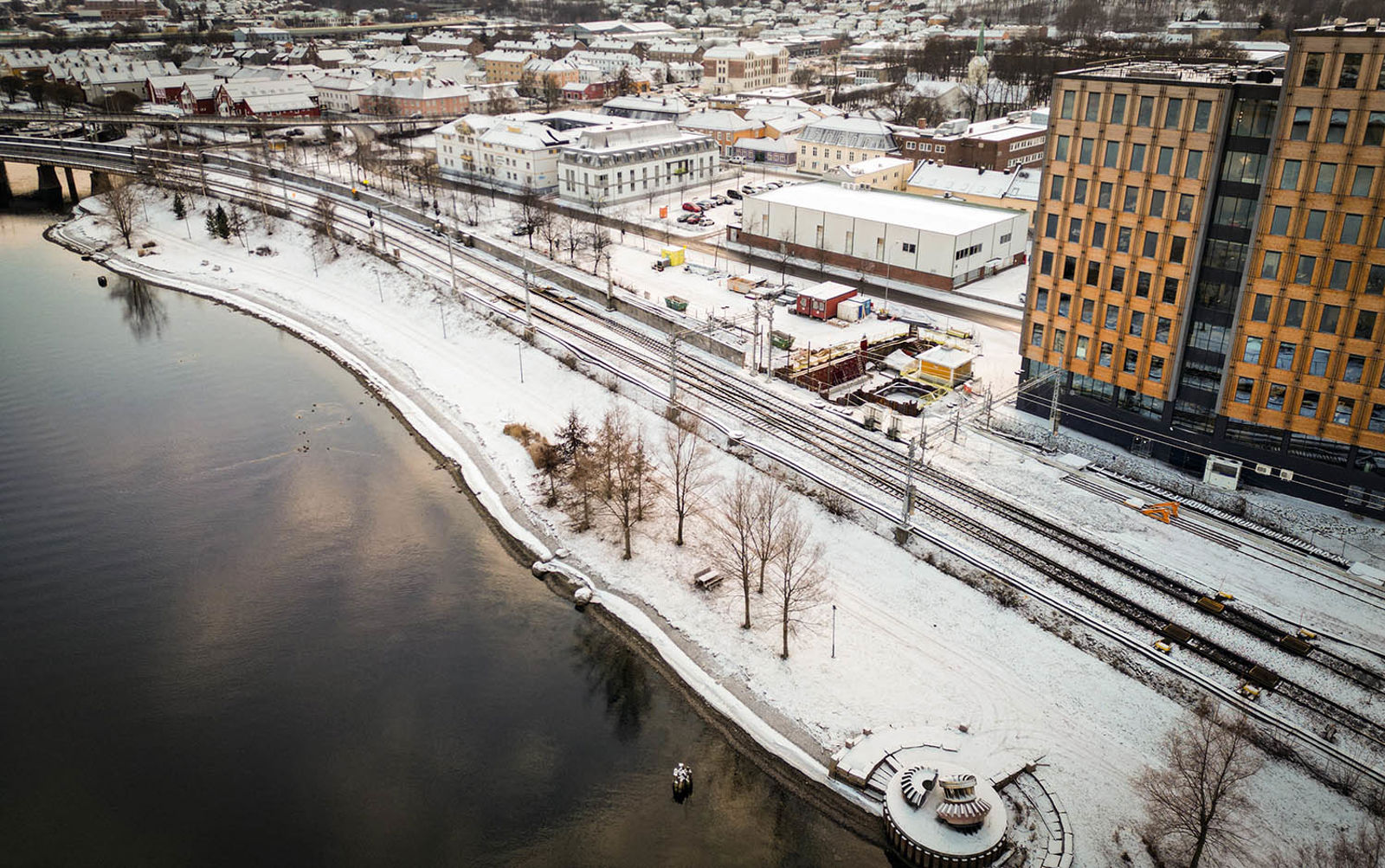 Sporområde og elvepromenade sett fra luften. Snø på bakken. Synlig anleggsområdet langs sporene.