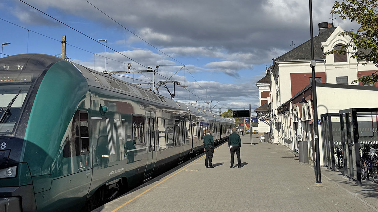 Grønt Vy-tog til venstre, personer på plattformen, stasjonsbygningen og sykkelparkering til høyre.